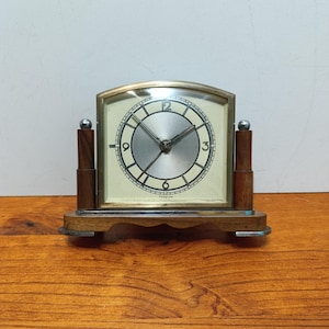 May include: A vintage table clock with a rectangular face and a gold-colored frame. The clock has a cream-colored face with black numerals and hands. The clock is supported by a wooden base and two wooden pillars with silver-colored ball accents.
