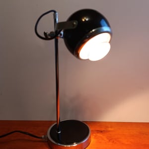 Puede incluir: Una lámpara de escritorio negra y cromada con una base redonda y un cabezal esférico. El cabezal de la lámpara está inclinado hacia abajo, emitiendo una luz brillante. La lámpara tiene un brazo curvo y un acabado cromado pulido.