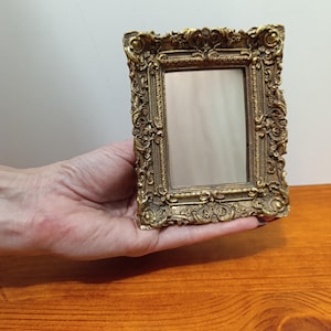 Puede incluir: Marco de fotos dorado de estilo antiguo con detalles intrincados. El marco rectangular tiene un pequeño espacio vacío para una foto o una obra de arte. El marco se sostiene en una mano, mostrando su tamaño y diseño.
