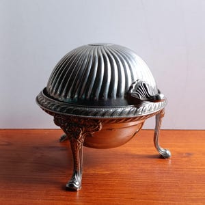 Puede incluir: Una mantequera antigua de color plateado con tapa abovedada y acanalada y patas curvas ornamentadas. El plato tiene un borde decorativo y un asa en la tapa. La mantequera se exhibe sobre una superficie de madera.