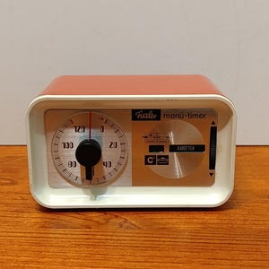 May include: A vintage Fissler menü-timer with a salmon-colored top and white frame. The timer features a circular dial with numbers and a black knob. The face includes the text "Fissler menü-timer" and "KAROTTEN".