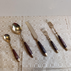 Può includere: Un set di cinque posate: due cucchiai, un coltello e due forchette. Le posate hanno teste in metallo dorato, manici in legno marrone scuro e dettagli in metallo dorato.