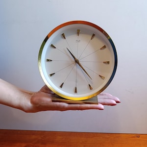 May include: A round analog table clock with a white face, gold-tone rim, and a square base. The clock features thin gold-tone hour markers, slender gold-tone hands, and the text "Kienzle Automatic" printed in black near the top center of the dial.
