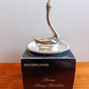 May include: A silver-plated swan ring holder featuring a swan figurine mounted on a circular, textured tray. The object sits atop a black box with the text "SILVERPLATED" and "Swan Ring Holder" printed in white, along with a small "SP2002" label.