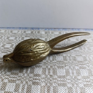 Könnte beinhalten: Ein Vintage-Nussknacker aus Messing mit detailliertem, strukturiertem Design. Der Nussknacker hat die Form eines stilisierten Vogels oder einer Frucht mit einem Scharniermechanismus zum Knacken von Nüssen. Die Griffe erstrecken sich vom Körper.
