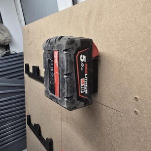 Op de afbeelding: Een zwarte en rode Milwaukee REDLITHIUM M18 batterij van 5,0 Ah is gemonteerd aan een muur met een zwarte metalen beugel.