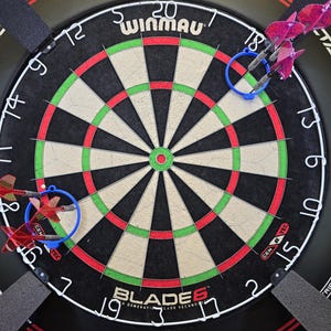 May include: A Winmau Blade 6 dartboard, black with red and green accents. The board has numbers around the edge and a central bullseye. Three darts with pink flights are in the board. The text "WINMAU" is at the top.