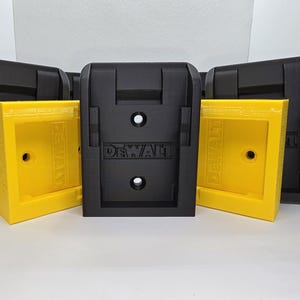 May include: Four black and yellow wall mounts for Dewalt batteries. The mounts have a rectangular shape with a recessed area for the battery to fit into. The mounts are labelled "DEWALT" in white letters.