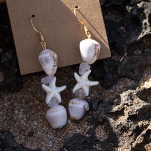 Könnte beinhalten: Ein Paar Ohrhänger mit natürlichen Muscheln, Seesternen und kleinen, hellvioletten Steinen. Die Ohrringe haben goldfarbene Haken. Die Muscheln sind weiß und beige, und die Seesterne sind weiß. Die Ohrringe werden vor einem braunen Hintergrund präsentiert.