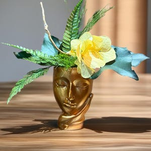 Könnte beinhalten: Eine goldfarbene Gesichtsvase mit geschlossenen Augen und einer Hand, die das Kinn stützt. Die Vase ist mit grünem Laub und einer gelben Blume gefüllt. Die Anordnung befindet sich auf einer Holzoberfläche.