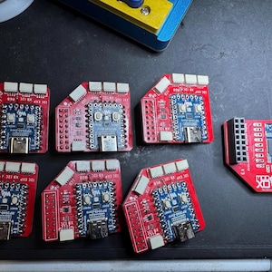 May include: A collection of red and blue circuit boards with various components, including micro USB ports and microcontrollers. Some boards have the text "X8 I2C EXP RGB" and "RP2040-Zero". The boards are arranged on a dark surface.