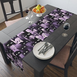 Könnte beinhalten: Ein Tischläufer mit einem sich wiederholenden Muster aus schwarzen Katzenköpfen auf lila Hintergrund. Der Läufer liegt auf einem dunklen Holztisch, mit einer weißen Obstschale, Weingläsern und Tellern mit Gabeln.