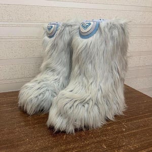 May include: A pair of light blue, furry winter boots. The boots have a high shaft and a round toe. Each boot has a patch with a heart-shaped logo in blue, white, and silver. The boots are made of a soft, shaggy material.