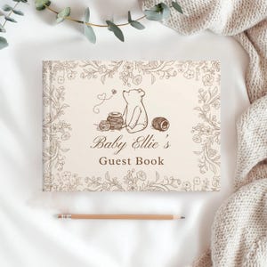 May include: A rectangular guest book with a cream-colored cover featuring a brown line-art illustration of a bear sitting near honey pots, surrounded by a floral border. The text reads Baby Ellie's Guest Book in a brown script and serif font.