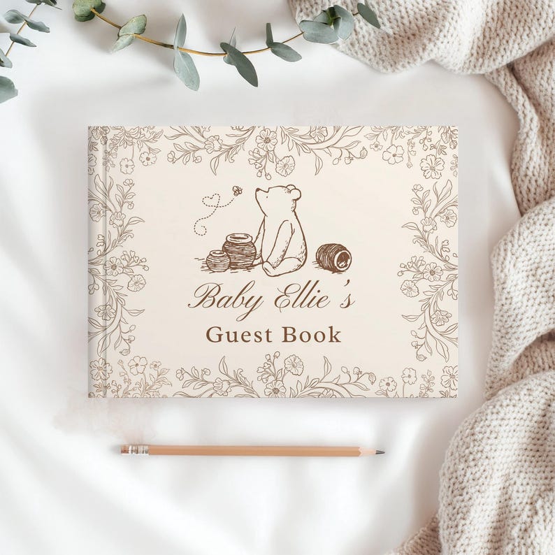 May include: A rectangular guest book with a cream-colored cover featuring a brown line-art illustration of a bear sitting near honey pots, surrounded by a floral border. The text reads Baby Ellie's Guest Book in a brown script and serif font.