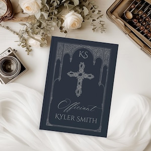 May include: A navy blue book with a decorative silver cross and the word "Officiant" in script. The book cover also features the initials "KS" and the name "Kyler Smith". The book is surrounded by flowers and a vintage camera.