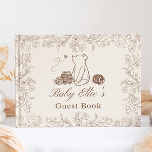 May include: A rectangular guest book with a cream-colored cover featuring a brown line-art illustration of a bear sitting near honey pots, surrounded by a floral border. The text reads Baby Ellie's Guest Book in a brown script and serif font.