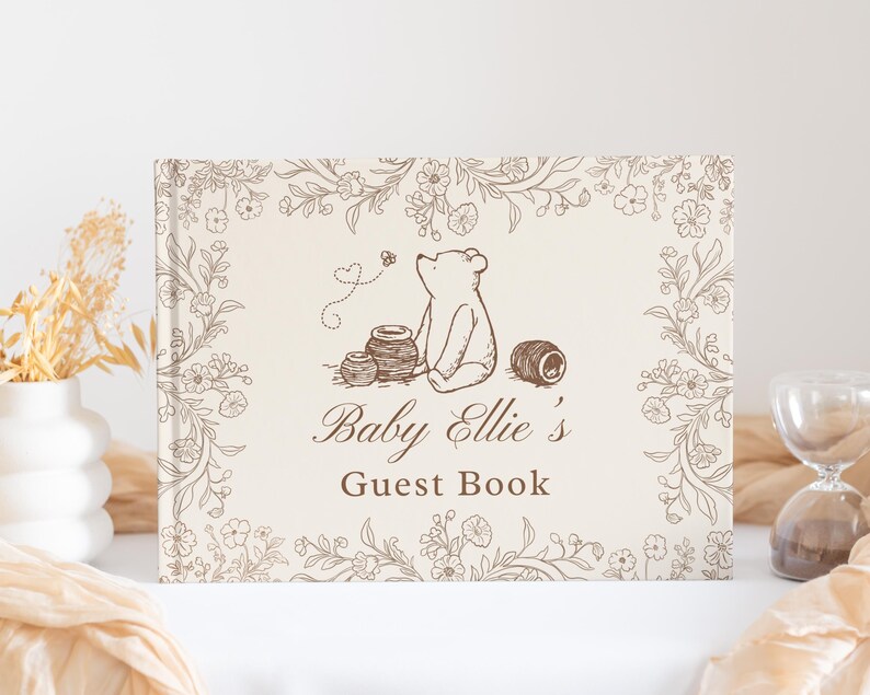 May include: A rectangular guest book with a cream-colored cover featuring a brown line-art illustration of a bear sitting near honey pots, surrounded by a floral border. The text reads Baby Ellie's Guest Book in a brown script and serif font.