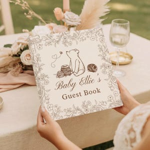 May include: A square guest book with a cream-colored cover featuring a brown line-art illustration of a bear, honey pots, and a bee. The text reads Baby Ellie's Guest Book, surrounded by a delicate floral border in a matching brown ink.