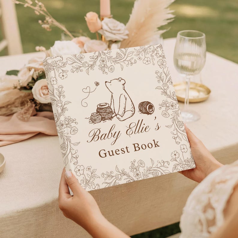 May include: A square guest book with a cream-colored cover featuring a brown line-art illustration of a bear, honey pots, and a bee. The text reads Baby Ellie's Guest Book, surrounded by a delicate floral border in a matching brown ink.