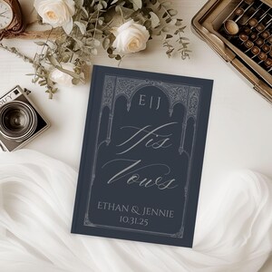 May include: A navy blue "His Vows" book with ornate silver lettering and a decorative border. The book includes the names Ethan & Jennie and the date 10.31.25. The image is styled with flowers, a vintage camera, and a typewriter.