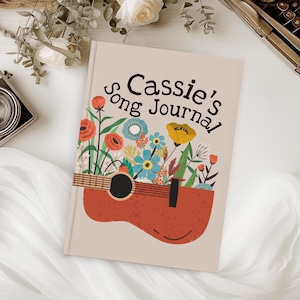 May include: A beige journal with the text "Cassie's Song Journal" in black lettering. The journal is decorated with a red acoustic guitar with a floral design. The flowers are red, yellow, blue, and green.
