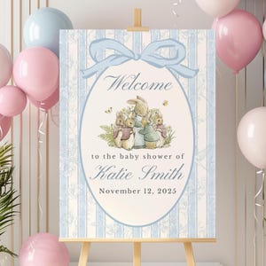 May include: A baby shower welcome sign on an easel. The sign features a blue bow and the word "Welcome" in script. Below, it reads "to the baby shower of Katie Smith, November 12, 2025." The sign has a floral and striped background with illustrations of bunnies.