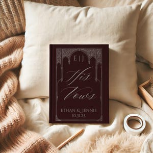 May include: A burgundy-colored book titled "His Vows" with white script and decorative border. The book includes the names Ethan & Jennie and the date 10.31.25. The book is resting on a cream-colored pillow and blanket.