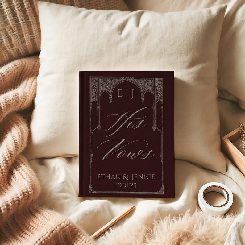 May include: A burgundy-colored book titled "His Vows" with white script and decorative border. The book includes the names Ethan & Jennie and the date 10.31.25. The book is resting on a cream-colored pillow and blanket.