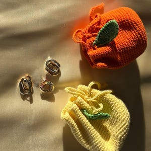 Könnte beinhalten: Zwei kleine gehäkelte Taschen in Form von Früchten. Eine Tasche ist orange und die andere ist gelb. Beide Taschen haben ein grünes Blattdetail.