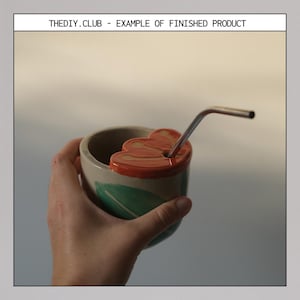 May include: A handmade ceramic cup with a flower-shaped lid and a stainless steel straw. The cup is beige with a green leaf design and an orange rim. The text "THEDIY.CLUB - EXAMPLE OF FINISHED PRODUCT" is at the top.