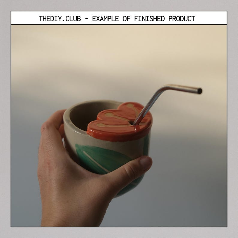 May include: A handmade ceramic cup with a flower-shaped lid and a stainless steel straw. The cup is beige with a green leaf design and an orange rim. The text "THEDIY.CLUB - EXAMPLE OF FINISHED PRODUCT" is at the top.