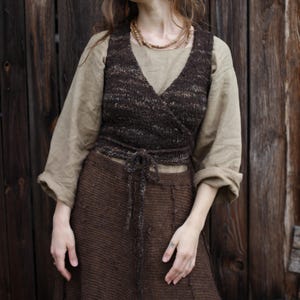 Könnte beinhalten: Ein braunes Strick-Wickeloberteil mit Bindeband, kombiniert mit einem passenden braunen Rock. Das Oberteil hat einen V-Ausschnitt und ist über einem hellbraunen Langarmshirt geschichtet. Das Outfit besteht aus Naturfasern und hat eine rustikale, böhmische Ästhetik.