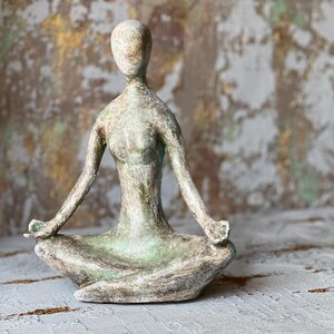 Puede incluir: Una escultura de una figura de yoga sentada en posici&oacute;n meditativa. La estatua es de color verde y gris apagado, con los brazos extendidos y las manos apoyadas en las rodillas. La escultura est&aacute; sobre una superficie texturizada.