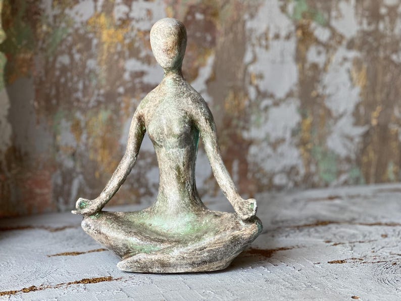Puede incluir: Una escultura de una figura de yoga sentada en posici&oacute;n meditativa. La estatua es de color verde y gris apagado, con los brazos extendidos y las manos apoyadas en las rodillas. La escultura est&aacute; sobre una superficie texturizada.