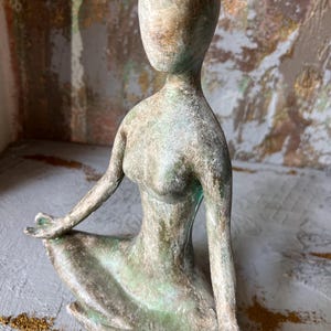 Puede incluir: Escultura de una figura sentada, sin rostro, en pose meditativa. La escultura tiene un aspecto envejecido con tonos verdes y marrones, sugiriendo una est&eacute;tica antigua. La figura est&aacute; sentada sobre una superficie gris, con un fondo texturizado.