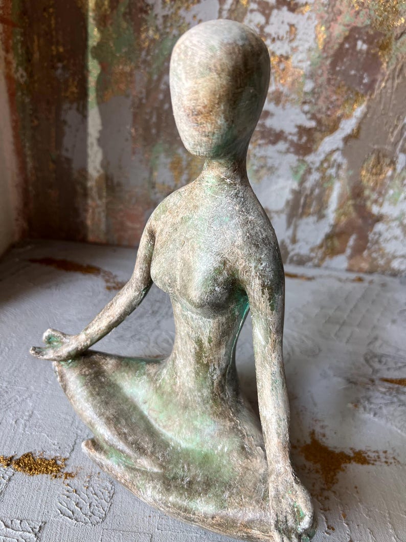 Puede incluir: Escultura de una figura sentada, sin rostro, en pose meditativa. La escultura tiene un aspecto envejecido con tonos verdes y marrones, sugiriendo una est&eacute;tica antigua. La figura est&aacute; sentada sobre una superficie gris, con un fondo texturizado.