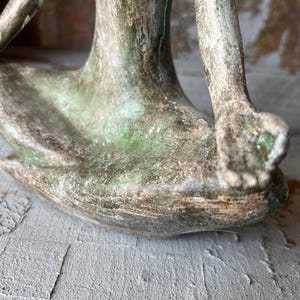 Puede incluir: Una escultura de bronce de una figura sentada en una postura de yoga. La escultura tiene un aspecto envejecido con tonos verdes y marrones. Los brazos y las piernas de la figura son visibles, con las manos apoyadas en las rodillas. La escultura est&aacute; sobre una superficie gris.