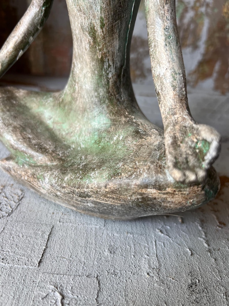 Puede incluir: Una escultura de bronce de una figura sentada en una postura de yoga. La escultura tiene un aspecto envejecido con tonos verdes y marrones. Los brazos y las piernas de la figura son visibles, con las manos apoyadas en las rodillas. La escultura est&aacute; sobre una superficie gris.
