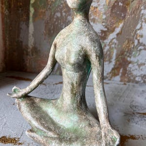 Puede incluir: Una escultura de color bronce de una figura sentada en posici&oacute;n meditativa. La figura tiene una forma lisa y alargada, con los brazos apoyados en las rodillas y las manos en un gesto de paz. La escultura tiene un aspecto envejecido.