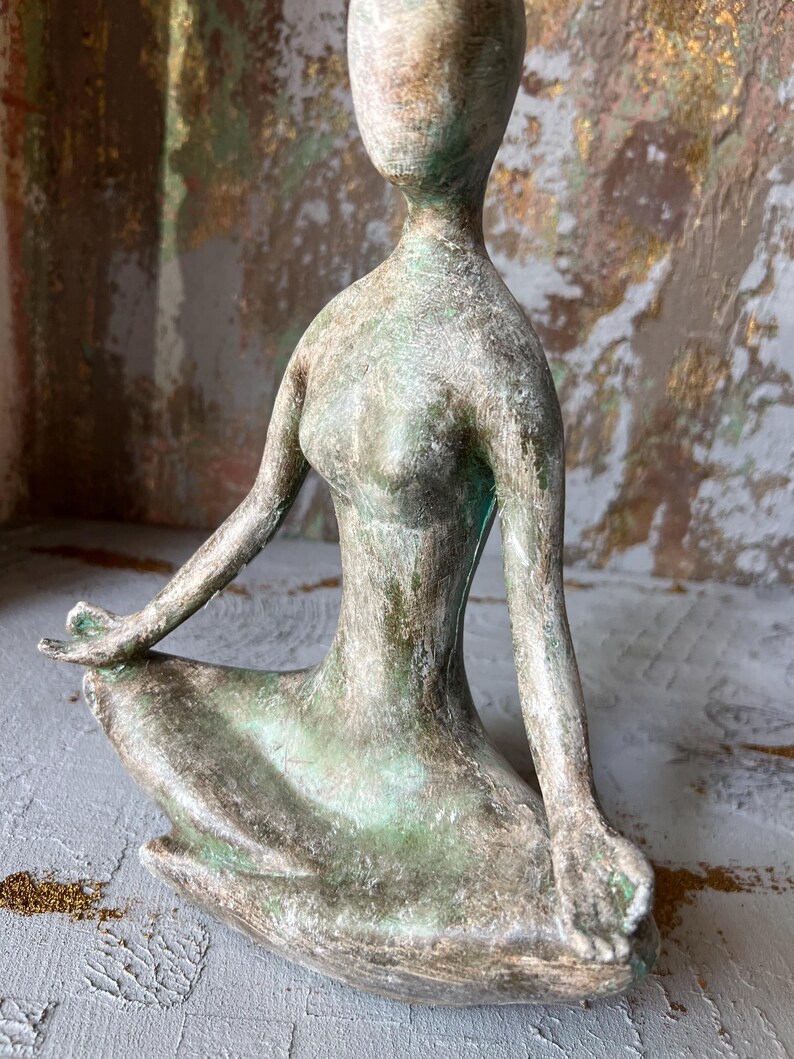 Puede incluir: Una escultura de color bronce de una figura sentada en posici&oacute;n meditativa. La figura tiene una forma lisa y alargada, con los brazos apoyados en las rodillas y las manos en un gesto de paz. La escultura tiene un aspecto envejecido.