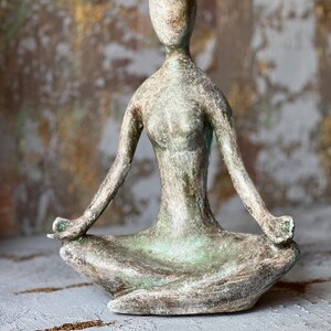 Puede incluir: Una escultura de tono bronce envejecido de una persona en una postura de yoga sentada. La figura tiene los brazos extendidos, las manos descansando sobre las rodillas y est&aacute; en una posici&oacute;n meditativa. La escultura tiene una superficie texturizada y una expresi&oacute;n serena.