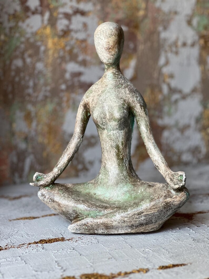 Puede incluir: Una escultura de tono bronce envejecido de una persona en una postura de yoga sentada. La figura tiene los brazos extendidos, las manos descansando sobre las rodillas y est&aacute; en una posici&oacute;n meditativa. La escultura tiene una superficie texturizada y una expresi&oacute;n serena.
