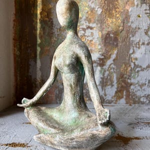 Puede incluir: Una escultura de figura sentada y desgastada, en posici&oacute;n meditativa. La escultura tiene una paleta de colores apagados de verde, gris y bronce. Los brazos de la figura est&aacute;n extendidos y las manos descansan en el regazo. Es un objeto decorativo.