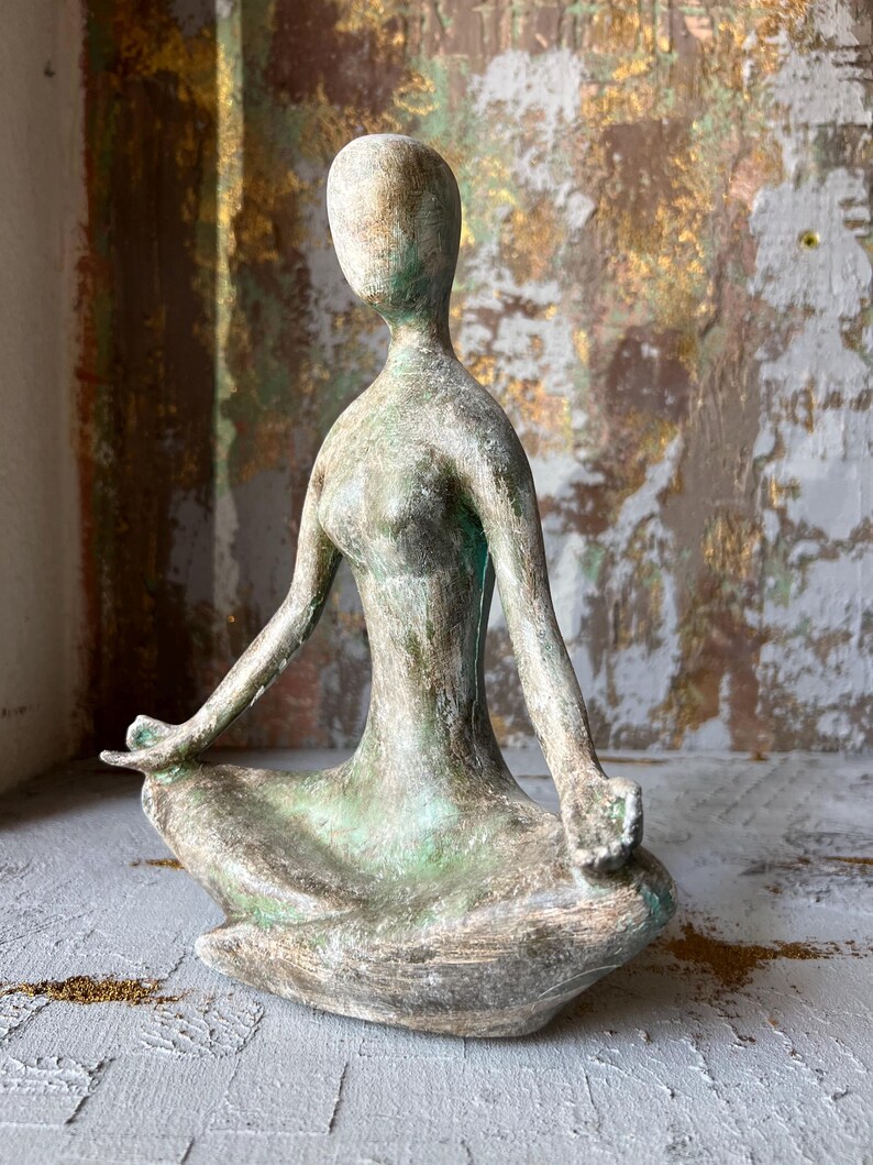 Puede incluir: Una escultura de figura sentada y desgastada, en posici&oacute;n meditativa. La escultura tiene una paleta de colores apagados de verde, gris y bronce. Los brazos de la figura est&aacute;n extendidos y las manos descansan en el regazo. Es un objeto decorativo.
