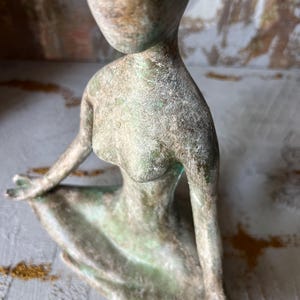 Puede incluir: Una escultura de bronce de una figura sentada con rasgos alargados. La figura tiene una cabeza y brazos lisos y redondeados. La escultura tiene un aspecto envejecido y texturizado con toques de verde y marr&oacute;n.