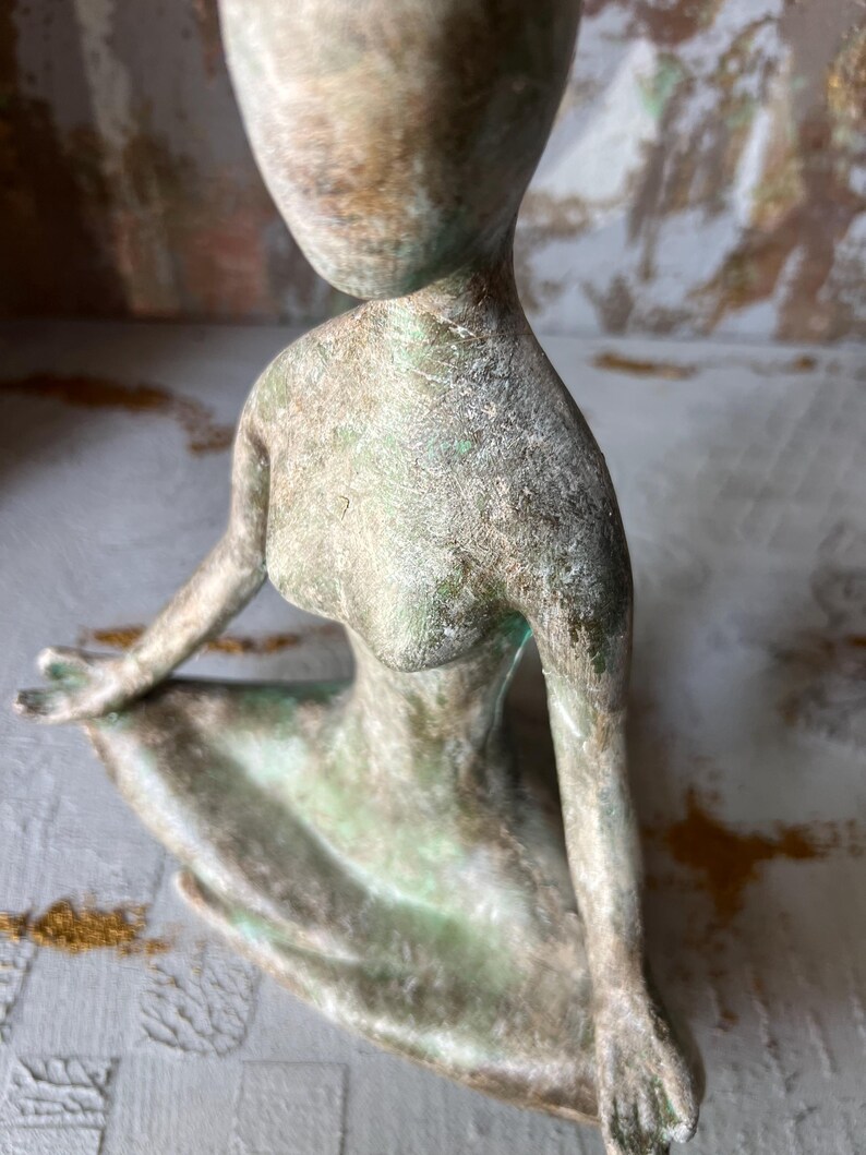 Puede incluir: Una escultura de bronce de una figura sentada con rasgos alargados. La figura tiene una cabeza y brazos lisos y redondeados. La escultura tiene un aspecto envejecido y texturizado con toques de verde y marr&oacute;n.
