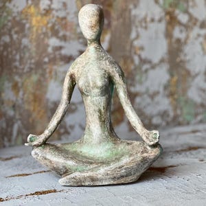 Puede incluir: Escultura de una persona en postura de yoga sentada. La figura est&aacute; en posici&oacute;n de loto con los brazos extendidos y las manos en un mudra. La escultura tiene una paleta de colores verde y gris apagados, lo que sugiere una apariencia natural y envejecida.
