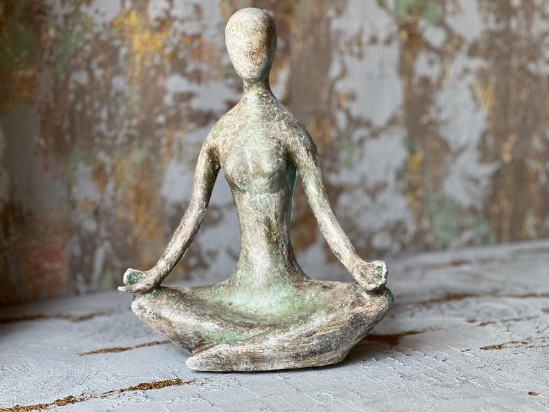 Puede incluir: Escultura de una persona en postura de yoga sentada. La figura est&aacute; en posici&oacute;n de loto con los brazos extendidos y las manos en un mudra. La escultura tiene una paleta de colores verde y gris apagados, lo que sugiere una apariencia natural y envejecida.