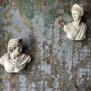 May include: Two plaster busts of people with detailed facial features and hair. The busts are mounted on a textured, gray wall with gold and green accents.